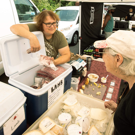 Two women at a vendor tent displaying meats and  cheeses